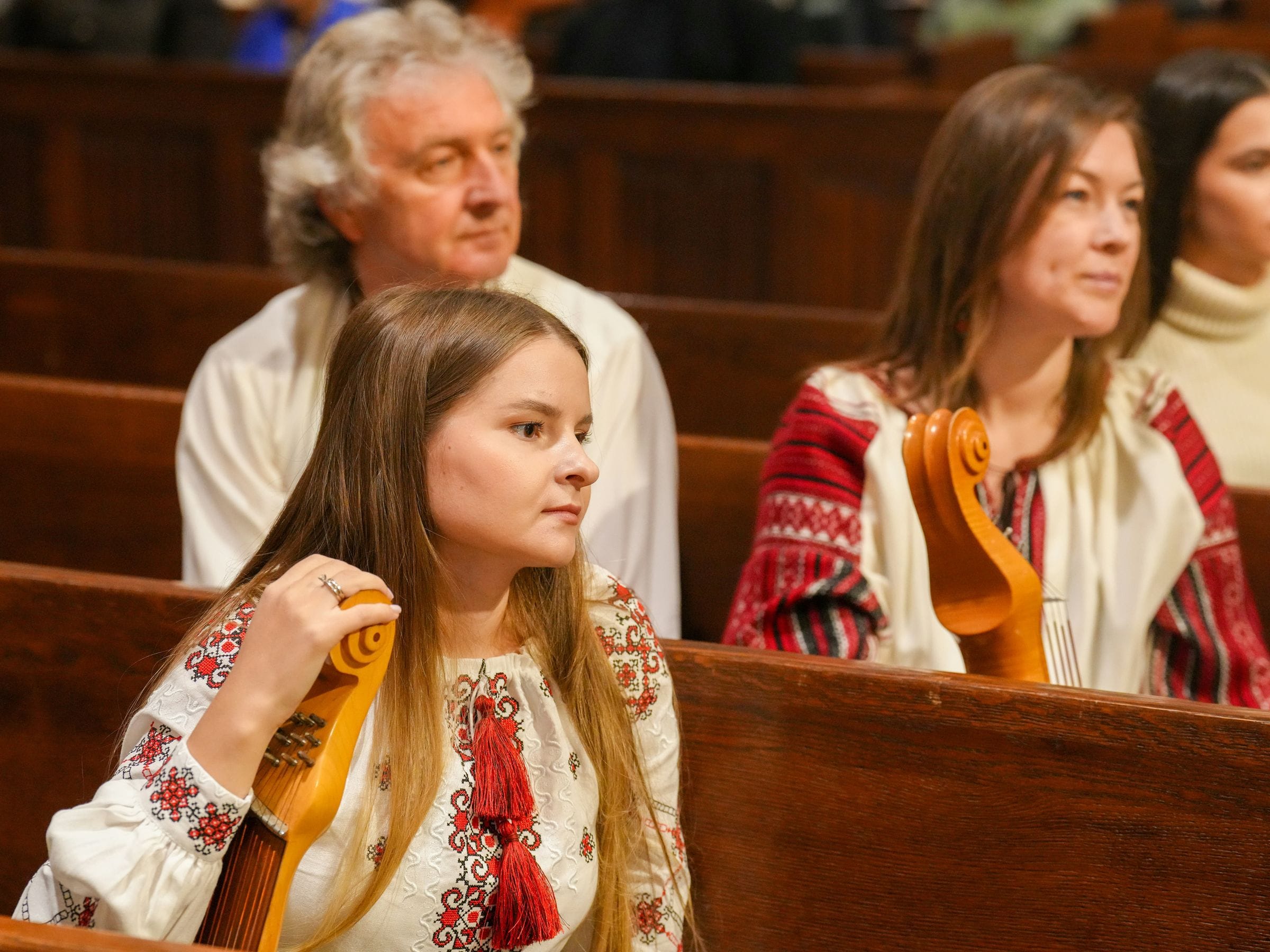 Músicos con banduras y camisas ucranianas bordadas sentados en bancos de madera de una iglesia.