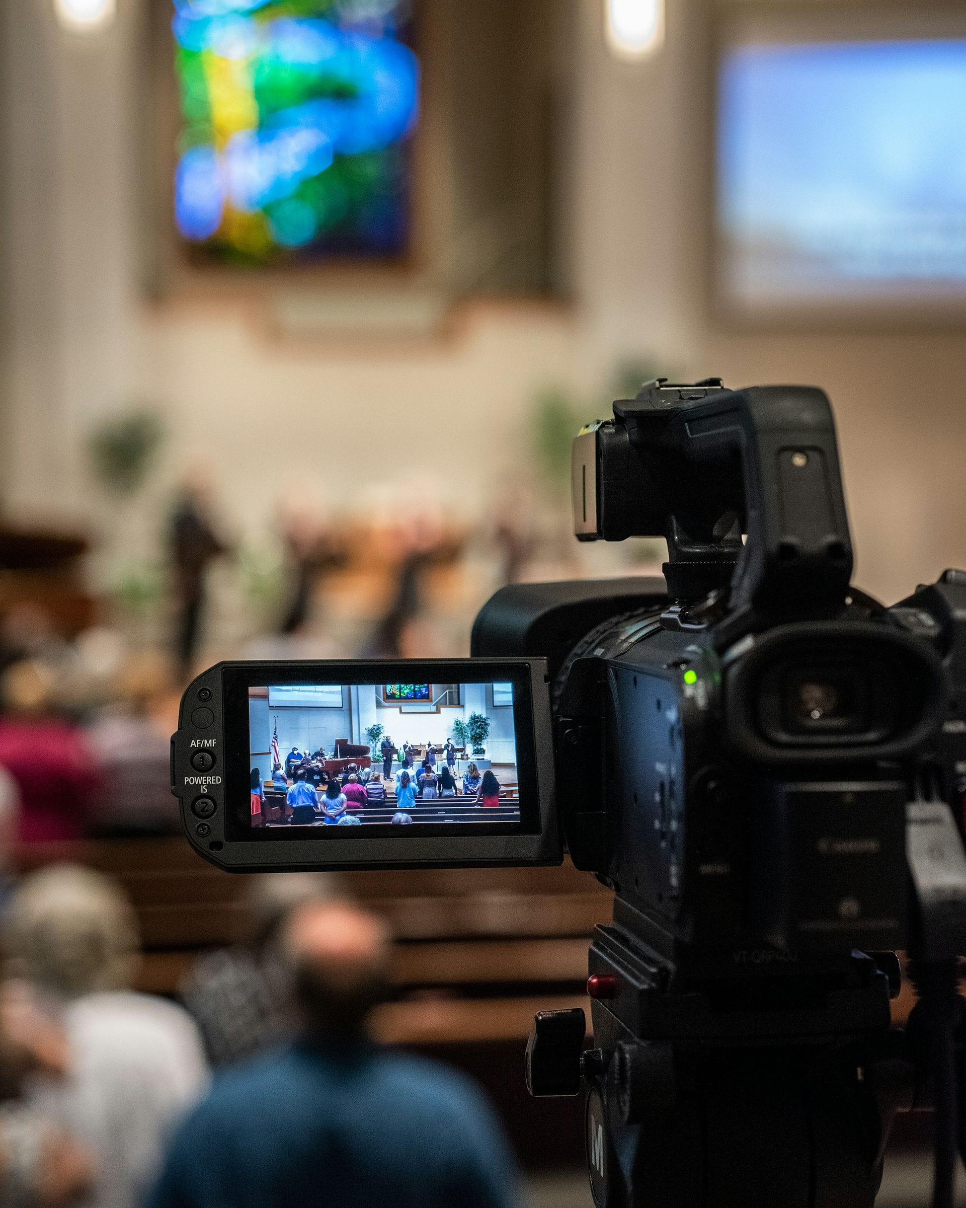 Una cámara de video graba un servicio; su pantalla muestra la congregación y el escenario.